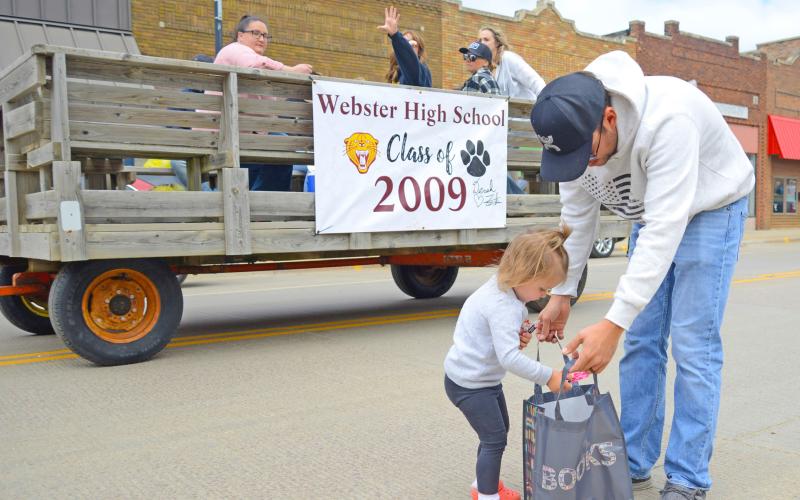 Two-year-old Cora Geier drops fistfuls of candy into a waiting bag held open by her dad Jadon as members of the Webster High School class of 2009 pass by in the background, tossing more sweet treats her direction during the Webster alumni parade June 29 on Main Street in Webster. Cora’s mom is Mariah (Block) Geier who is a member of the 2014 Webster Area High School graduating class. Reporter &amp;amp; Farmer | Amanda Dulitz