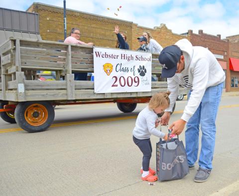Two-year-old Cora Geier drops fistfuls of candy into a waiting bag held open by her dad Jadon as members of the Webster High School class of 2009 pass by in the background, tossing more sweet treats her direction during the Webster alumni parade June 29 on Main Street in Webster. Cora’s mom is Mariah (Block) Geier who is a member of the 2014 Webster Area High School graduating class. Reporter &amp; Farmer | Amanda Dulitz