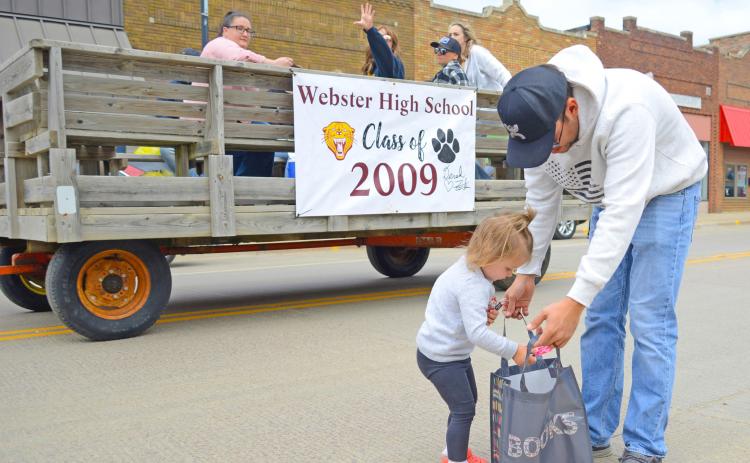 Two-year-old Cora Geier drops fistfuls of candy into a waiting bag held open by her dad Jadon as members of the Webster High School class of 2009 pass by in the background, tossing more sweet treats her direction during the Webster alumni parade June 29 on Main Street in Webster. Cora’s mom is Mariah (Block) Geier who is a member of the 2014 Webster Area High School graduating class. Reporter &amp; Farmer | Amanda Dulitz